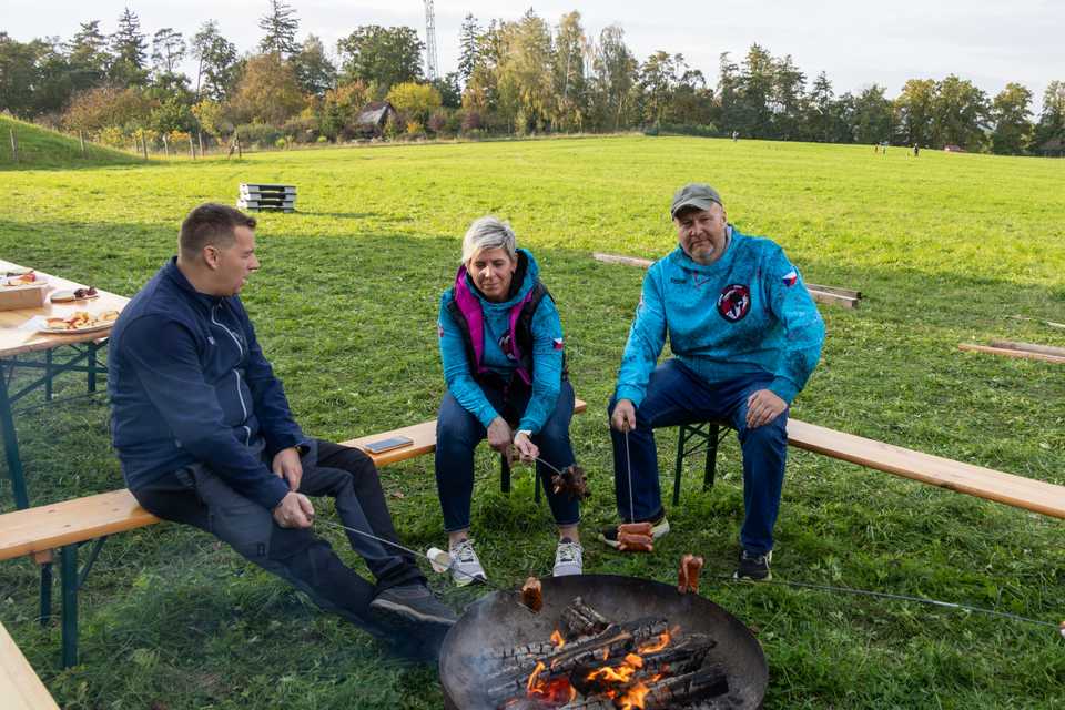 Tři dospělí sedí u ohniště a opékají špekáčky.